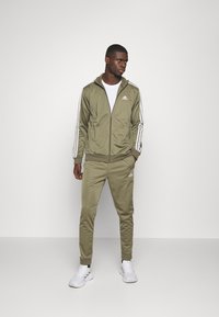 Olivovo zelený teplákový súprava vyrobená z hladkého materiálu, s bielymi pruhmi na rukávoch a nohách, so zipsom na bundě a elastickými manžetami.