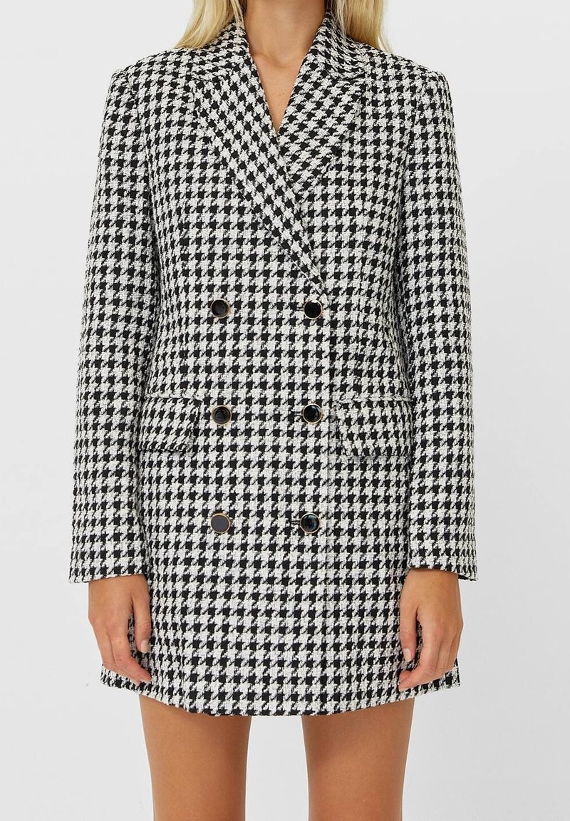 Femme portant une robe blazer à double boutonnage noir et blanc à motif pied-de-poule avec des boutons noirs, debout devant un fond uni.