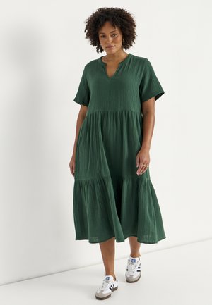 Nainen, jolla on päällään vihreä teksturoitu midimekko lyhyillä hihoilla ja valkoiset tennarit mustilla raidoilla, seisoo valkoista taustaa vasten.