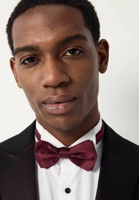 Un smoking noir avec un nœud papillon en satin bordeaux, une chemise blanche et un accent de col assorti. Peau lisse et cheveux coupés de près, mettant l'accent sur le visage.