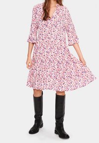 Květinové šaty v růžové barvě s fialovými a červenými vzory, výstřih do V, rukávy 3/4 a rozšířený lem. Černé kozačky do kolen dotvářejí vzhled.