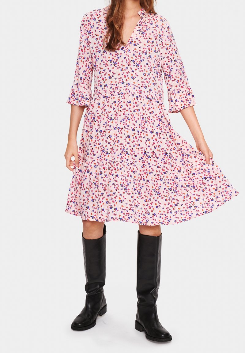 Květinové šaty v růžové barvě s fialovými a červenými vzory, výstřih do V, rukávy 3/4 a rozšířený lem. Černé kozačky do kolen dotvářejí vzhled.
