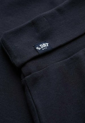 Donker marineblauwe gebreide stof met een klein aangebracht label waarop "S.DRY NYC" staat, vlakbij een gevouwen rand.