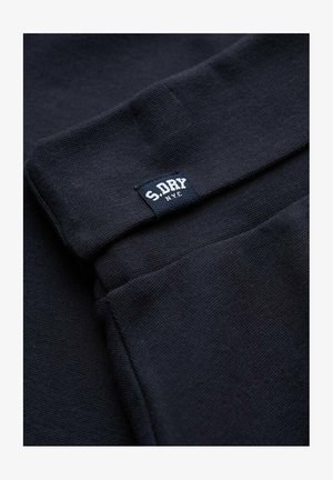 Tessuto a maglia blu navy scuro con una piccola etichetta cucita che dice "S.DRY NYC" vicino a un bordo piegato.