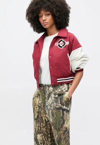 Μπορντό μπουφάν bomber με λευκά μανίκια και ριγωτές μανσέτες, διακοσμημένο με κυκλικό λογότυπο Jordan. Συνδυάζεται με παντελόνι καμουφλάζ με σχέδια.