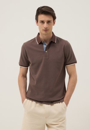 Joven con las manos en los bolsillos, vistiendo una camiseta polo marrón de manga corta con ribete blanco y pantalones beige, de pie frente a un fondo liso.