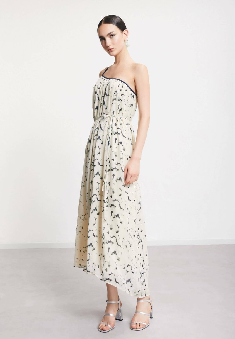 Femme portant une robe crème sans manches longue jusqu'aux chevilles avec des motifs abstraits noirs et des sandales argentées à talons hauts, debout sur un sol gris.