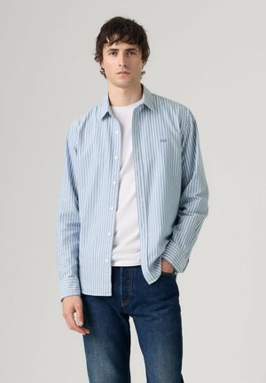 Junger Mann, der ein hellblau-weiß gestreiftes Hemd über einem weißen T-Shirt und dunkelblauen Jeans trägt, steht vor einem einfachen Hintergrund.