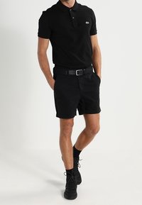 Homme portant un polo noir, un short noir ceinturé, des chaussettes noires et des baskets noires, se tenant avec les mains dans les poches.