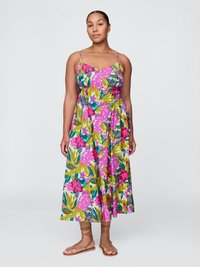 Frau trägt ein ärmelloses, knopfbares, tropisches Blumenkleid mit grünen, pinken und blauen Blättern und Blumen, kombiniert mit braunen Riemchensandalen.