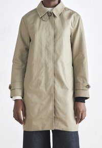 Imperméable kaki avec un col à boutons, des manches longues et des poches latérales. Présente une texture lisse et une légère brillance.