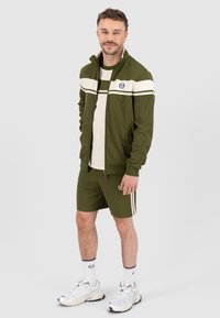 Olivgrünes Sportoutfit bestehend aus einer Jacke mit Reißverschluss und Shorts, mit weißen Streifen und Logo. Inklusive weißer Sneakers und gestreifter Socken.