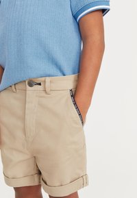 Beige shorts met opgerolde omslagen, voorzien van een metalen knoopsluiting en een zwart accent op de zijzak, gecombineerd met een blauwe geblokte shirt.