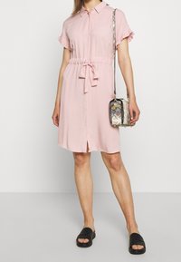 Rosa kortærmet kjole med krave og flæsedetaljer, indsnævet talje og knaplukning foran; kombineret med en mønstret crossbody-taske og sorte sandaler.