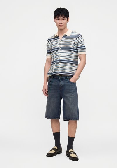 Homme debout, une main dans la poche, portant une chemise à manches courtes rayée, un short en jean foncé, des chaussettes noires et des mocassins bicolores.