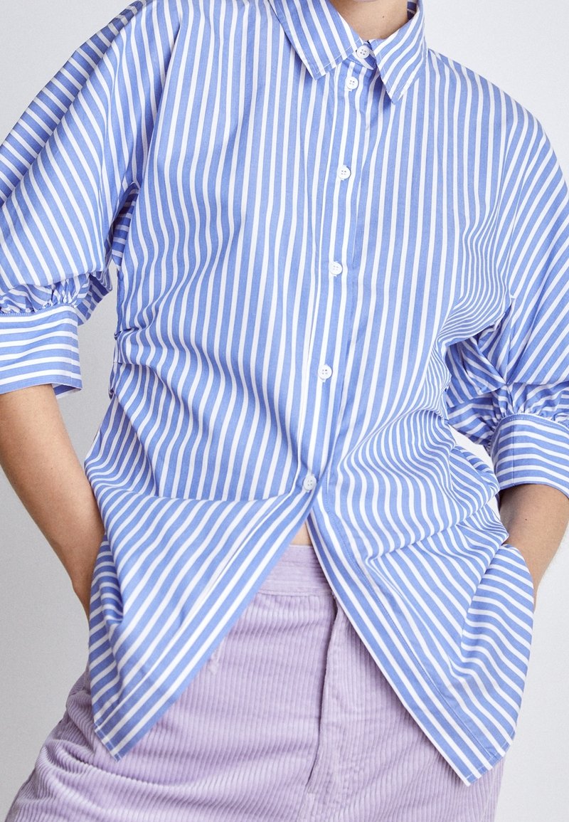 Personne portant une chemise à rayures verticales bleues et blanches à boutons, avec des manches froncées trois-quarts, et un pantalon en velours côtelé violet clair.