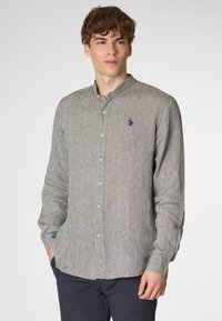 Giovane con capelli ricci che indossa una camicia grigia con bottoni, si trova contro uno sfondo neutro, con le mani nelle tasche, guardando direttamente l'osservatore.