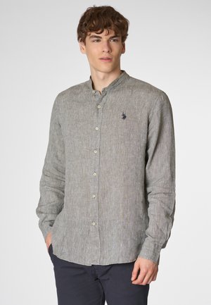 Giovane con capelli ricci che indossa una camicia grigia con bottoni, si trova contro uno sfondo neutro, con le mani nelle tasche, guardando direttamente l'osservatore.