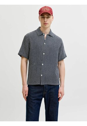 Jeune homme portant une chemise à manches courtes à motifs, un jean foncé et une casquette de baseball rouge avec un texte brodé.