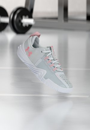 Chaussure de sport gris clair et rose suspendue en l'air sur le sol de la salle de sport, avec un haltère flou en arrière-plan.