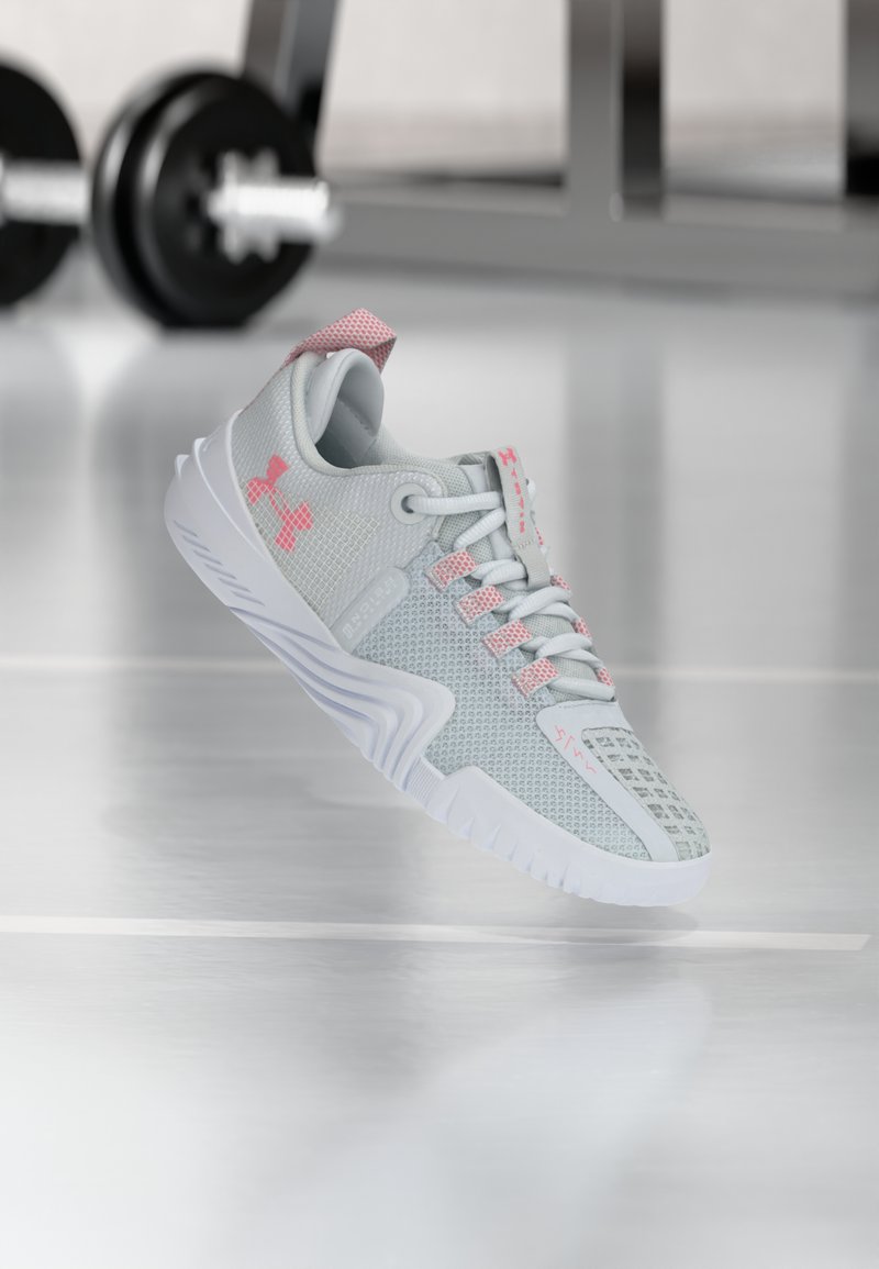 Αθλητικό παπούτσι ανοιχτού γκρι και ροζ αιωρείται στον αέρα πάνω σε γυμναστήριο, με θολό αλτήρα στο παρασκήνιο.