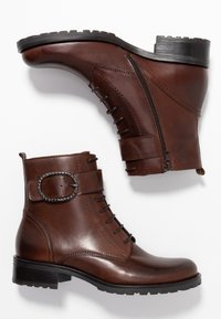 Bottes courtes en cuir marron avec une fermeture éclair latérale, un design à lacets et une bande décorative avec une boucle ovale. Elles possèdent une semelle texturée.