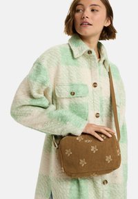 Braune Cord-Tasche mit floralem Stickdesign, flaches Design, verstellbarer Tragegurt und subtilen goldenen Beschlägen. Kombination mit einer grün karierten Jacke.