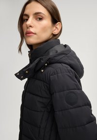 Zwarte donsjas met een glanzende afwerking, voorzien van een ritssluiting aan de voorkant, een capuchon met trekkoord en gestikte details. Logo op de linkermouw.