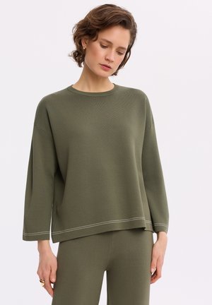 Kobieta ubrana w oliwkowo-zieloną luźną bluzkę z długim rękawem i dopasowane spodnie, patrząca w dół na jednolity jasny tle.