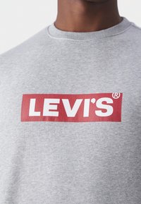 Homme portant un sweat-shirt gris clair à col rond avec un logo Levi's rouge rectangulaire centré sur la poitrine.
