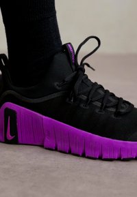 Pantofilor sport negri cu un superior texturat, având o talpă din cauciuc violet și logo-ul Nike. Șireturile sunt negre, iar interiorul are un guler căptușit.