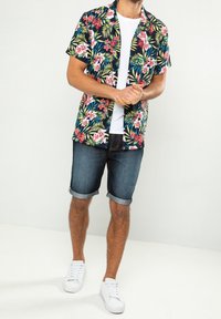 Hombre vestido con una camisa de flores de manga corta en azul marino, una camiseta blanca, pantalones cortos de mezclilla arremangados y zapatillas blancas, de pie contra un fondo claro y liso.