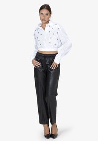Vrouw die met handen op de heupen staat, gekleed in een witte cropped blouse met sterrenpatronen, zwarte high-waisted leren broek en zwarte hakken.
