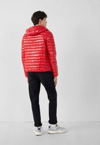 Giacca a vento rossa con finitura lucida, caratterizzata da un motivo trapuntato orizzontale e un cappuccio, abbinata a pantaloni neri e sneakers chiare.