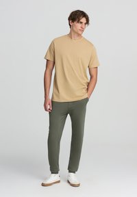 Maglietta beige a maniche corte abbinata a pantaloni jogger verde oliva e sneakers bianche. L'outfit presenta una vestibilità comoda e una texture di tessuto morbido.