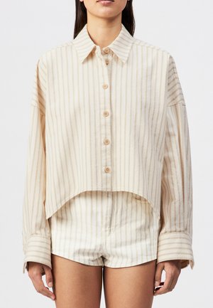 Vrouw draagt een beige-wit gestreept overhemd met knopen en bijpassende korte broek, staand tegen een effen achtergrond.