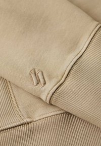 Tessuto di felpa beige con polsini a costine; presenta un piccolo logo ricamato. Texture liscia con dettagli di cucitura visibili.