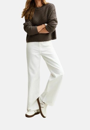 Personne portant un pull brun texturé, un pantalon large blanc, des baskets blanches aux semelles marron, les bras croisés, et aux cheveux longs.