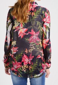 Blouse fleurie en matériau léger et transparent avec un fond sombre et des motifs de fleurs tropicales dans des tons rose vif, jaune et vert.