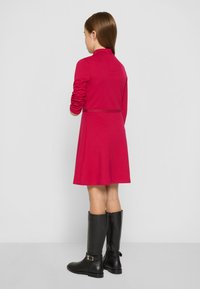 Piros, hosszú ujjú ruha gallérral, csillogó derékpánttal, fekete térdcsizmával, amelyen csat részlet található.