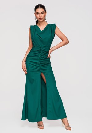 Kobieta w bezrękawnikowej szmaragdowej sukience z marszczeniami i wysokim rozcięciem na udzie, nosząca srebrne szpilki, kolczyki koła i bransoletki, pozująca na jednolitym tle.