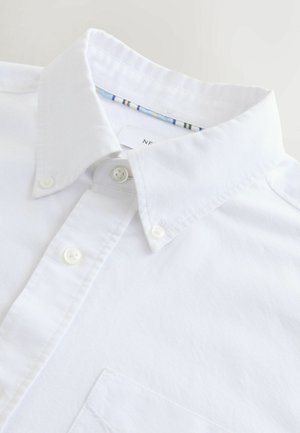 Weißes Hemd mit Knöpfen, mit einem zugeknöpften Kragen und einer Brusttasche, flach auf einer hellen Oberfläche ausgelegt.