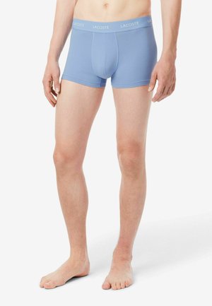 Staande persoon die lichtblauwe Lacoste boxershorts draagt, getoond van taille tot voeten, op een witte achtergrond.