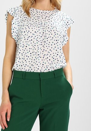 Vrouw draagt een witte korte mouw blouse met blauwe en groene polkadots en hoog getailleerde donker groene broek met handen in de zakken.