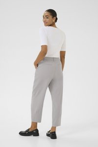 Femme en chemise blanche à manches courtes, pantalon gris clair ajusté et mocassins noirs, se tenant les mains dans les poches, regardant par-dessus son épaule.