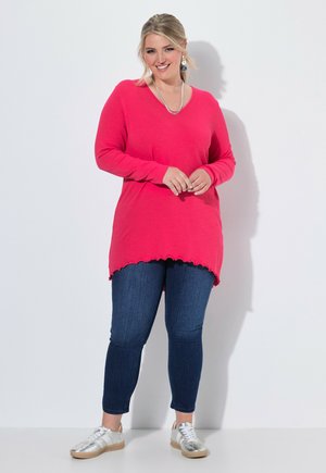 Vrouw staand in een felroze longsleeve, donkerblauwe spijkerbroek en zilveren sneakers tegen een effen witte achtergrond.