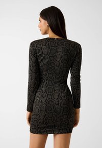 Kobieta o długich, ciemnych włosach nosi dopasowaną, długorękawową mini sukienkę w czarno-szary wzór wężowy, stoi tyłem na jednolitym szarym tle.