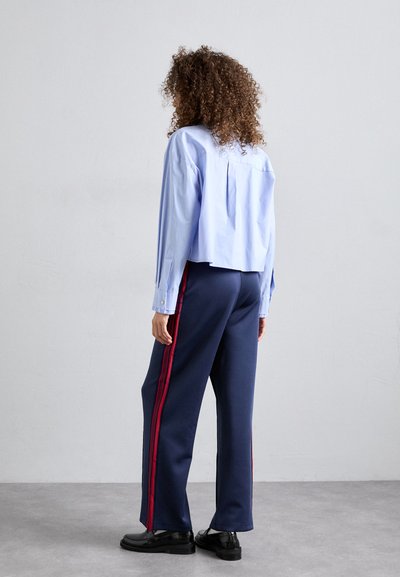 Mujer con cabello rizado que lleva una camiseta corta azul claro, pantalones navy de pierna ancha con rayas rojas y mocasines negros, de pie sobre un suelo gris.