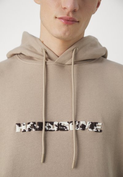 Sudadera beige con bolsillo tipo canguro, cordones y "HANDSOME" bordado en negro y blanco, con un detalle texturizado y con patrón.