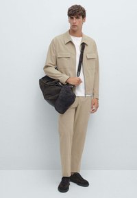 Jeune homme portant une veste et un pantalon beiges avec une chemise blanche, tenant un sac à bandoulière noir et une casquette, se tenant devant un fond clair uni.
